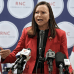 Florida Attorney General Ashley Moody in front of an RNC background wearing a red jacket with media microphones held up to her.