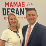 A blond woman thrusting out her chest is wearing a white short with blue trim. She stands next to a white man in a suit with a red tie. The sign behind them reads Mamas for DeSantis.