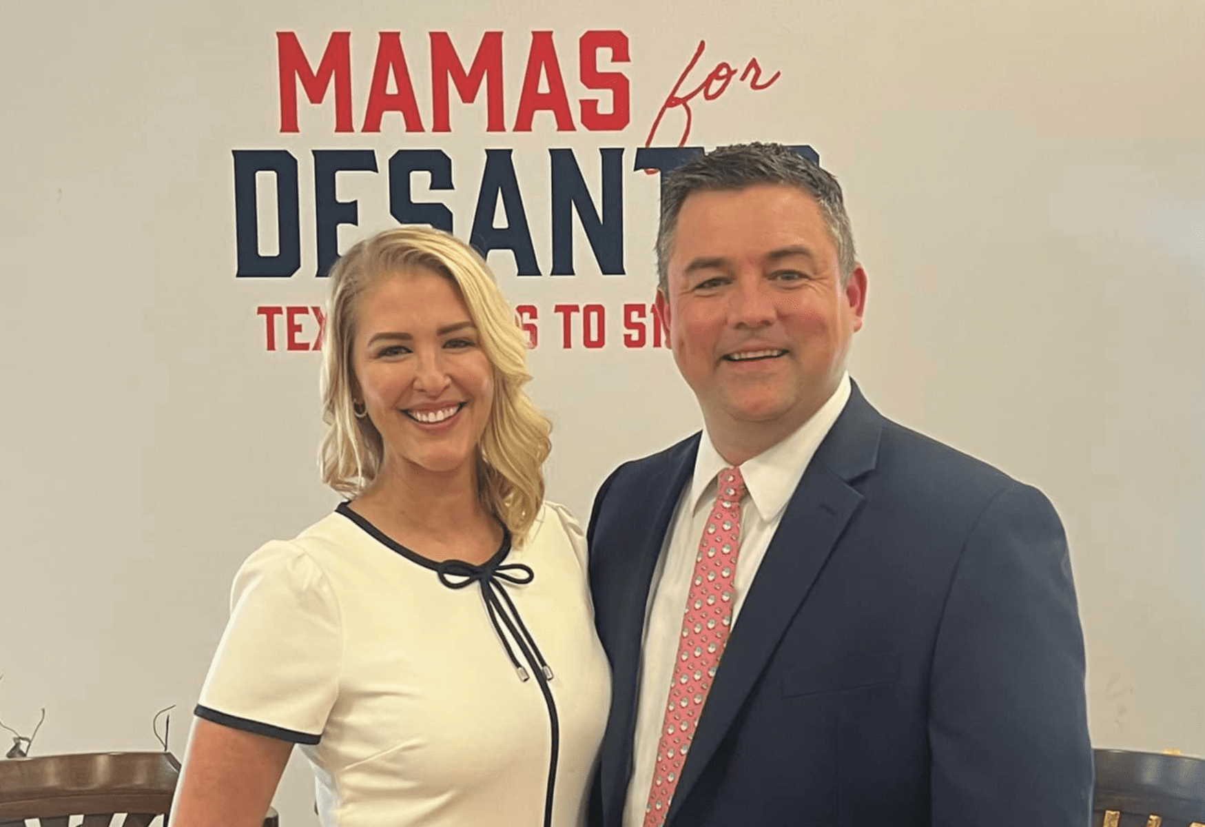 A blond woman thrusting out her chest is wearing a white short with blue trim. She stands next to a white man in a suit with a red tie. The sign behind them reads Mamas for DeSantis.
