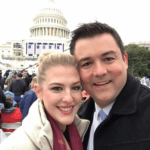 A snuggling smiling couple in front of a crowd in from of the Capitol. A blond woman in a white coat, a white man with black hair in a black coat.