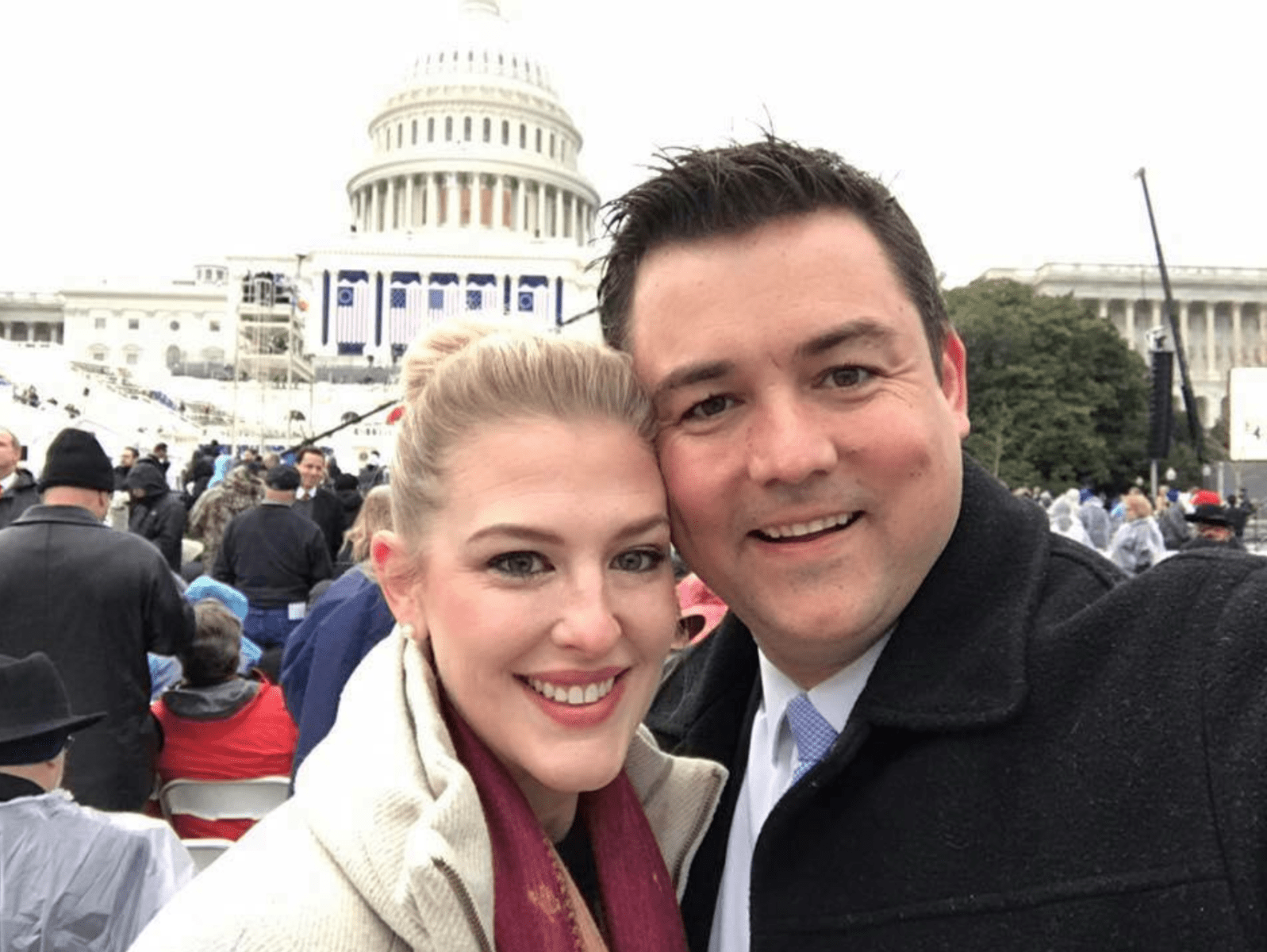 A snuggling smiling couple in front of a crowd in from of the Capitol. A blond woman in a white coat, a white man with black hair in a black coat.