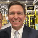 Three people in a warehouse with a sign that says Group Leaders. Left to right: a white man with a buzz cut, a white man with a grin (Ron DeSantis, a blond woman.