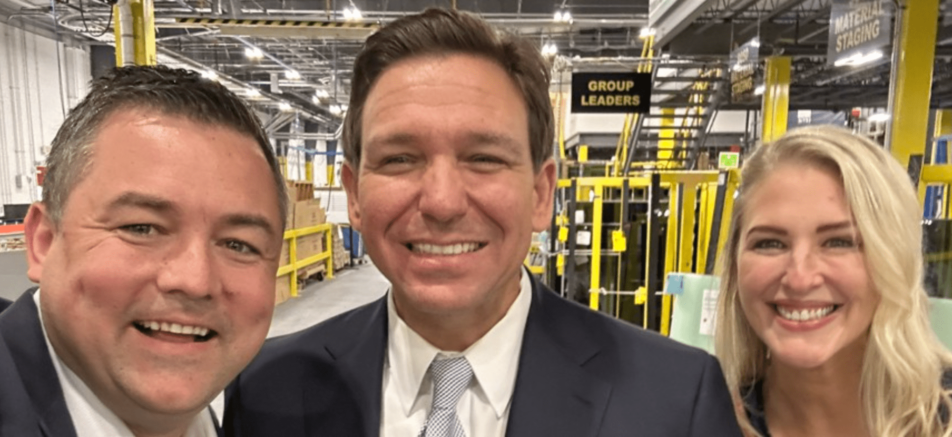 Three people in a warehouse with a sign that says Group Leaders. Left to right: a white man with a buzz cut, a white man with a grin (Ron DeSantis, a blond woman.